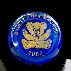 Puede incluir: Un pisapapeles redondo de vidrio azul con un diseño de oso de peluche dorado en el centro. El texto "Birth of A New Millennium" está alrededor del borde, con "2000" en la parte inferior. El pisapapeles está sobre una superficie negra.