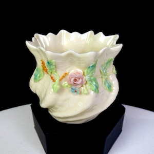 May include: A cream-colored porcelain vessel with a scalloped rim and a pearlescent finish. The exterior is decorated with three-dimensional ceramic floral accents, including a central pink rose, small blue flowers, and green leaves.