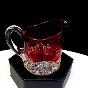 May include: A small ruby red glass creamer jug featuring a clear, diamond-patterned base and a curved handle. The upper portion of the red glass is etched with the name Bertha's in a cursive script. The jug has a pouring spout.