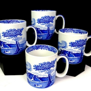 Puede incluir: Cuatro tazas de cerámica blanca con un diseño de paisaje azul. Cada taza presenta una ilustración detallada de un árbol, agua y figuras. Las tazas tienen un asa curva y un borde con dibujos azules.