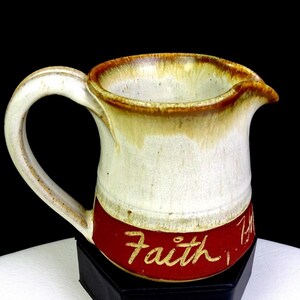 Puede incluir: Jarra de cerámica color crema con asa curva y pico vertedor. El borde y el asa tienen un esmalte marrón. Una banda roja en la base presenta la palabra "Faith" en escritura cursiva. La jarra se exhibe sobre un soporte negro.