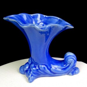 May include: A vibrant cobalt blue ceramic cornucopia vase featuring a wide, ruffled, trumpet-shaped opening and a curved, tapered base with decorative moulded scroll details. The glossy finish reflects light across the smooth, curved surface.