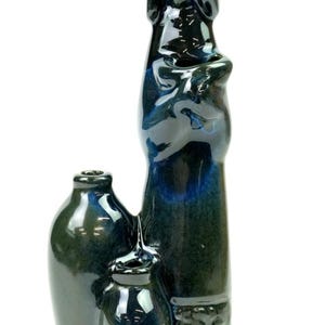 Może przedstawiać: Ciemnoniebieska i zielona szkliwiona rzeźba ceramiczna przedstawiająca stylizowaną postać stojącą obok dwóch małych, zaokrąglonych naczyń. Błyszczące wykończenie charakteryzuje się głębokimi odcieniami błękitu i leśnej zieleni z subtelnymi wariacjami kolorystycznymi.