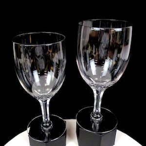 Puede incluir: Dos copas de vino transparentes con tallos largos. Las copas se exhiben sobre bases cuadradas negras sobre un fondo negro. Las copas están vacías y reflejan la luz.