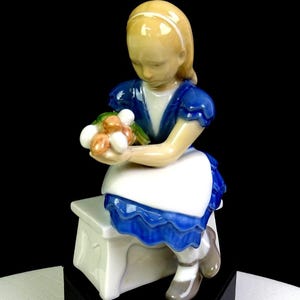May include: A porcelain figurine of a young girl seated on a white bench, wearing a blue dress with a white apron. She holds a bouquet of flowers. The figurine has blonde hair and is set against a black background.
