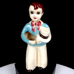 Puede incluir: Figura de cerámica de un niño con chaqueta azul claro, pantalón blanco y pajarita roja. Sostiene un sombrero blanco con una banda negra. La figura tiene el pelo rojo oscuro y está sobre una base negra.