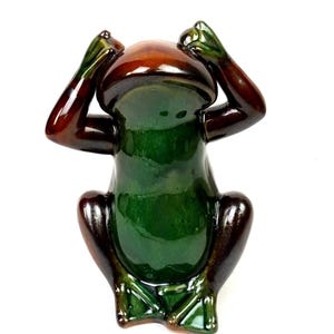 May include: A ceramic frog figurine in a seated position with its hands covering its eyes. The frog is glazed in glossy shades of green, brown, and dark brown. The figurine has a smooth, reflective surface and is set against a white background.