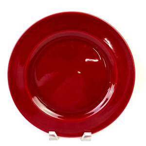 Puede incluir: Un plato llano circular con un acabado rojo sólido y brillante. El plato presenta un borde ancho y plano y un centro ligeramente hundido, mostrando una superficie lisa y reflectante en toda la pieza, visto desde una perspectiva cenital.