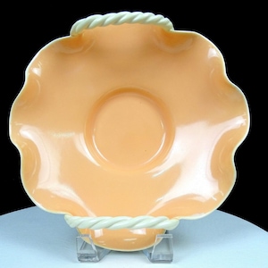 May include: A peach-coloured, scalloped-edge dish with a white rope-like trim. The dish has a circular indentation in the centre and sits on a clear plastic stand. The dish is likely made of glass or ceramic.