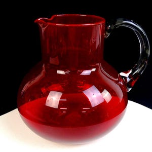 May include: A vibrant red glass pitcher with a rounded, bulbous body and a wide, open mouth featuring a small pouring spout. The pitcher includes a curved, dark-tinted glass handle attached to the side, providing a contrast to the deep red color.