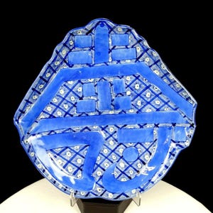 Puede incluir: Un plato decorativo de cerámica azul y blanco con una forma irregular. El plato presenta un patrón geométrico de líneas azules y detalles florales blancos. El diseño incluye una escritura azul estilizada. El plato se exhibe sobre un soporte negro.