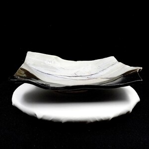 Könnte beinhalten: Eine quadratische Keramikschale mit weißem Innenraum und schwarzer Außenseite, die einen leicht geschwungenen, wellenförmigen Rand aufweist. Die Schale hat eine glatte, glänzende Oberfläche und steht auf einem runden weißen Sockel.
