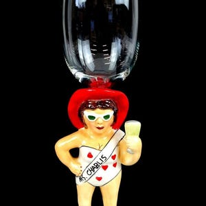 May include: A novelty wine glass featuring a ceramic figure of a woman wearing a red hat and holding a drink. The figure is attached to a clear glass. The woman is wearing a white swimsuit with red hearts and the text "Ms. Chablis".