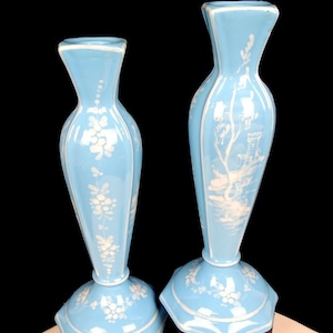 以下が含まれることがあります： 白の花柄と風景画が施された、水色のガラス花瓶2つ。花瓶はフレアトップと六角形のベースが特徴です。花瓶の高さは約30cmです。