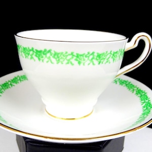 May include: White teacup and saucer set with a green floral design. The cup and saucer have gold trim. The cup has a curved handle. The saucer is round. The teacup and saucer are on a black background.