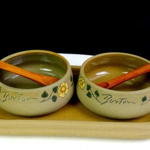 May include: Two round, light tan ceramic bowls with hand-painted yellow flowers and green leaves, each featuring the word Burton in script. The bowls sit on a matching rectangular tray with handles and include two wooden spoons for serving.