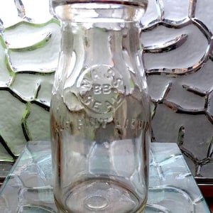 May include: Clear glass milk bottle with a rounded body and a narrow neck. The bottle has embossed text and a circular emblem. The words "PURE FIRST LIQUID" are visible. The bottle is sitting on a glass surface.