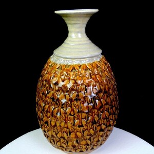 May include: A ceramic vase with a bulbous body featuring a textured, repeating leaf-like pattern in shades of brown and amber. The neck of the vase is smooth, flared at the top, and finished in a light cream glaze that contrasts with the dark body.