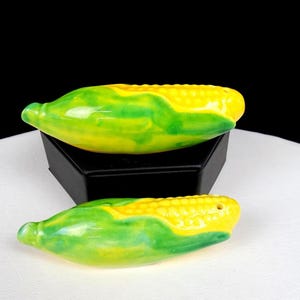 May include: A pair of ceramic salt and pepper shakers shaped like ears of corn. Each shaker features a bright yellow kernel section and green husks with a glossy finish. One shaker is positioned on a black stand, while the other rests below.