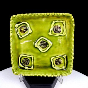 May include: A square ceramic plate with a glossy lime green glaze and a beaded edge. The surface features five raised, square-shaped indentations, each containing a dark brown, detailed leaf imprint centered within the lime green field.