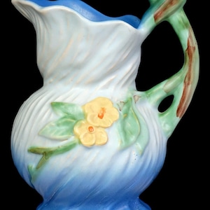 Puede incluir: Una jarra de cerámica con un acabado degradado en blanco y azul con una textura acanalada y en espiral. La jarra está adornada con un motivo floral amarillo en relieve con hojas verdes. Tiene un asa verde con forma de rama.