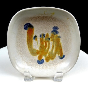 May include: A square ceramic dish with rounded corners, featuring a cream-colored base with a fine crackle glaze finish. The center is decorated with an abstract, hand-painted design in vibrant orange and deep blue, creating a textured pattern.
