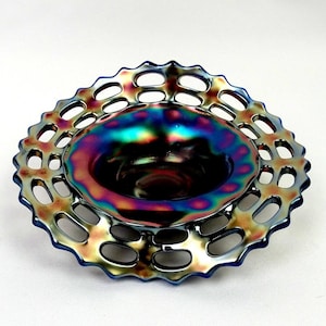 Könnte beinhalten: Eine schimmernde Glasschale mit gewelltem Rand und offenem Design. Die Schale hat eine tiefe, abgerundete Mitte und einen Rand mit einer Reihe ovaler Öffnungen. Die Farben wechseln von Blau über Lila zu Gold und erzeugen einen schimmernden Effekt.