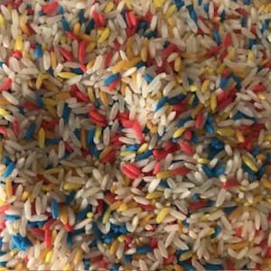 May include: Close-up of a pile of white rice grains mixed with colorful, dyed rice. The dyed rice is in shades of red, yellow, and blue, creating a vibrant, textured pattern. The image is well-lit, showing the details of each grain.