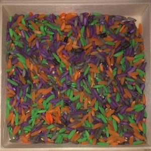 Puede incluir: Una bandeja cuadrada de madera llena de arroz colorido. El arroz está teñido en tonos naranja, verde y morado, creando una apariencia vibrante y texturizada. Los granos están dispersos por toda la bandeja.