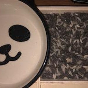 May include: A white bowl with a panda face design next to a wooden box filled with black and white rice. The bowl has a black rim and the panda face features black eyes and a nose.
