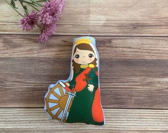 May include: A plush toy depicting a female saint with a crown, holding a sword and a palm branch. She is wearing a green dress with a red cape and a wheel behind her.