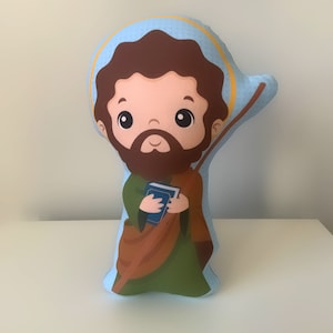 May include: A blue and brown plush pillow with a cartoon image of a man with a beard holding a book. The pillow is shaped like a person and has a halo around the head.
