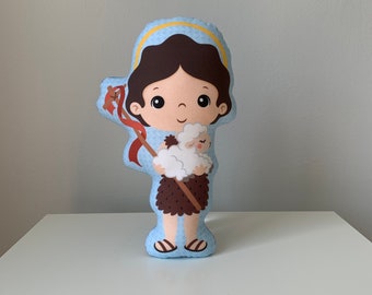 May include: A blue and brown plush toy of a shepherd carrying a white lamb. The shepherd has brown hair and a halo. The toy is a religious themed decoration.