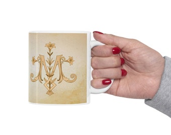 May include: White ceramic mug with a gold and beige design of a stylized "M" with floral accents. The mug has a handle and is held by a hand with red nail polish.