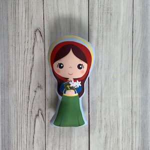 May include: A plush toy of a girl with brown hair, wearing a red dress and a yellow halo. She is holding a book and white flowers.