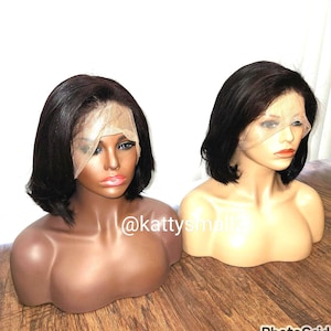 May include: Two mannequin heads displaying short, black wigs with lace fronts. The mannequins have tan skin tones and are positioned on a wooden surface. The text "@kattysmallz" is visible on the image.