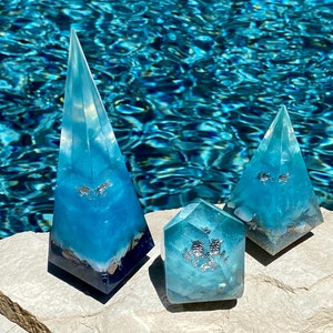 May include: Three blue resin pyramids with embedded seashells and silver glitter. The pyramids are different sizes and are sitting on a gray rock.