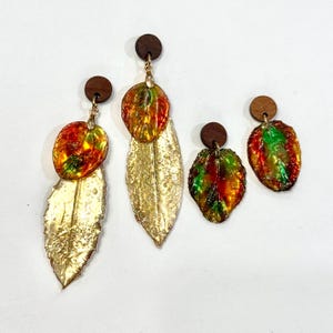 Puede incluir: Tres pares de pendientes de hoja de oro con hojas de resina de colores. Los pendientes son de diferentes tamaños y presentan una variedad de colores, incluyendo rojo, naranja, verde y amarillo.