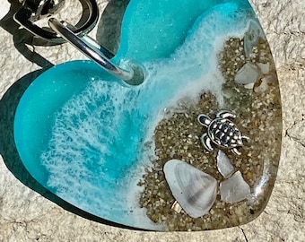 May include: A heart-shaped resin keychain with a blue ocean wave, white foam, and a small silver turtle on a sandy beach with seashells.