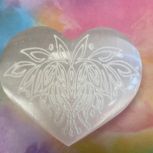 May include: White heart-shaped selenite crystal with a carved mandala design. The crystal is on a colorful background.