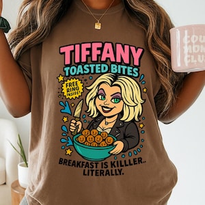 May include: Brown t-shirt featuring a cartoon graphic with the text "TIFFANY TOASTED BITES." The graphic includes a blonde woman holding a knife and a bowl of food, with the words "BREAKFAST IS KILLLER.. LITERALLY."