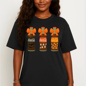 May include: Black t-shirt featuring three Coca-Cola bottle designs with Halloween themes. Each bottle has a different design: bats, ghosts, and candy corn. Orange bows adorn the bottle necks.