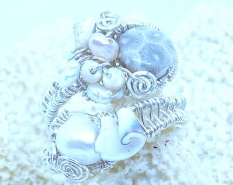 May include: A handcrafted ring featuring a seashell, pearls, and a coral stone, all intricately wrapped in silver wire. The ring's design incorporates spiral and woven patterns, creating a unique and elegant piece of jewelry.