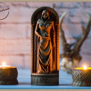 May include: A wooden statue of a woman in a hooded cloak, holding a skull. The statue is carved with intricate details and stands on a wooden base. The base is inscribed with the word "Hel".