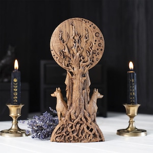 Op de afbeelding: Een bruine houten sculptuur met een boom, een ontwerp van een volle maan, twee wolfsfiguren en ingewikkelde gravures. De sculptuur staat op een wit oppervlak, met zwarte kaarsen in messing houders.