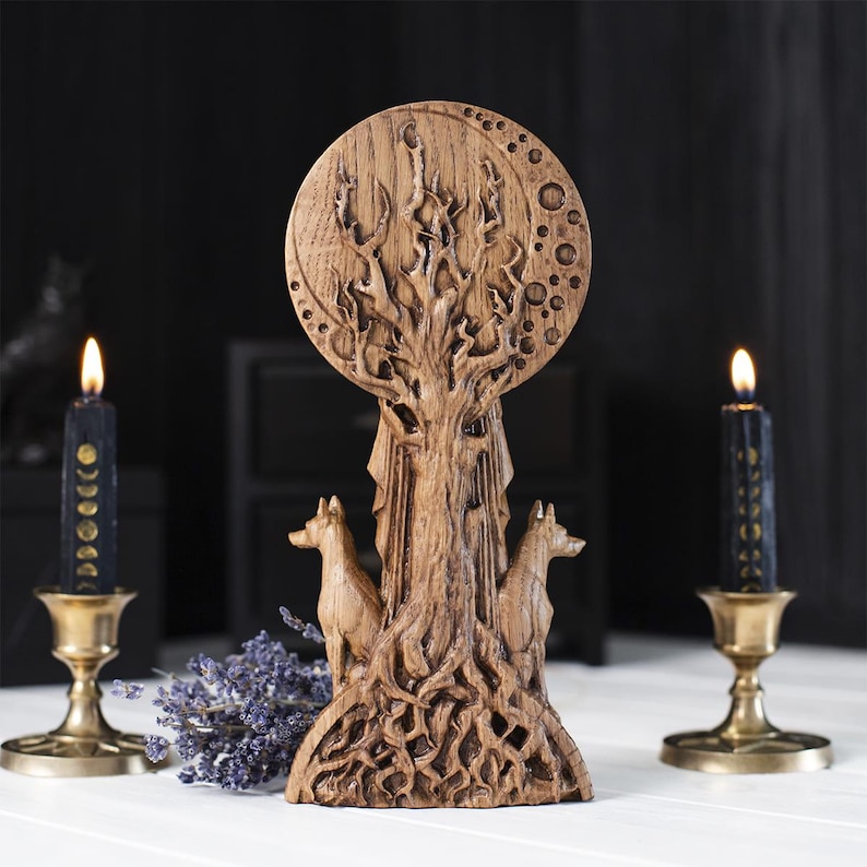 Op de afbeelding: Een bruine houten sculptuur met een boom, een ontwerp van een volle maan, twee wolfsfiguren en ingewikkelde gravures. De sculptuur staat op een wit oppervlak, met zwarte kaarsen in messing houders.