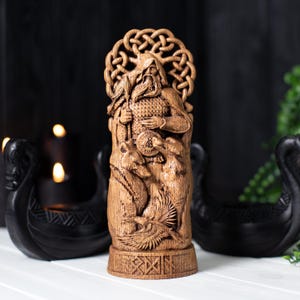 May include: A detailed wooden carving of a figure with a beard, holding a spear and shield, with two wolves and a bird at the base. The figure has a decorative knotwork halo. The carving is in shades of brown and stands on a base with carved symbols.