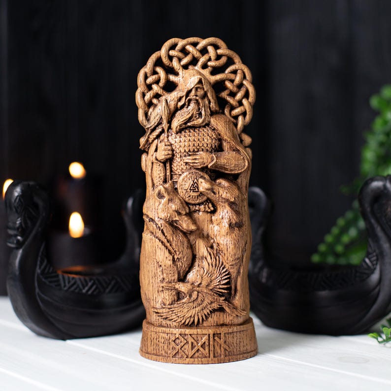 May include: A detailed wooden carving of a figure with a beard, holding a spear and shield, with two wolves and a bird at the base. The figure has a decorative knotwork halo. The carving is in shades of brown and stands on a base with carved symbols.
