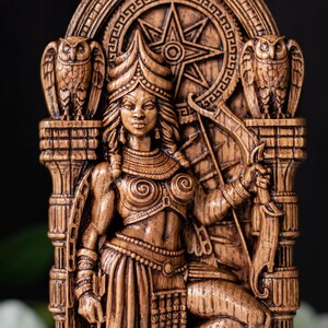 May include: A detailed wooden carving of a seated female figure, adorned with intricate jewelry and a headdress. Two owls flank the figure, and a starburst design is in the background. The figure holds a bow and arrow.