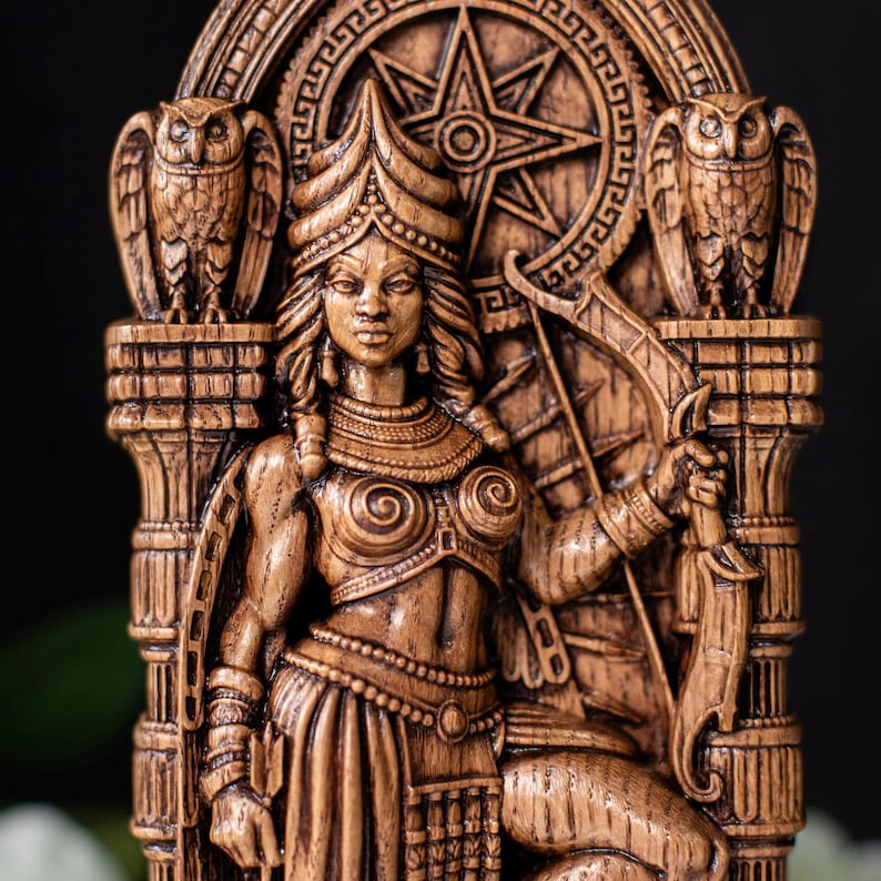 May include: A detailed wooden carving of a seated female figure, adorned with intricate jewelry and a headdress. Two owls flank the figure, and a starburst design is in the background. The figure holds a bow and arrow.